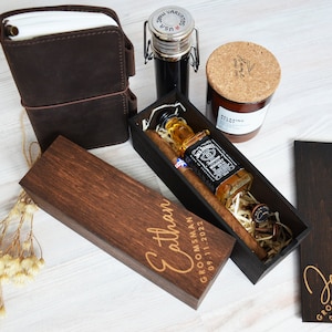 May include: A selection of gifts, including a brown leather journal, a small bottle with a metal clasp, a candle, and a wooden box containing a bottle of Jack Daniel's whiskey and a cigar. The box is engraved with the name "Ethan" and the date "09.11.2022".