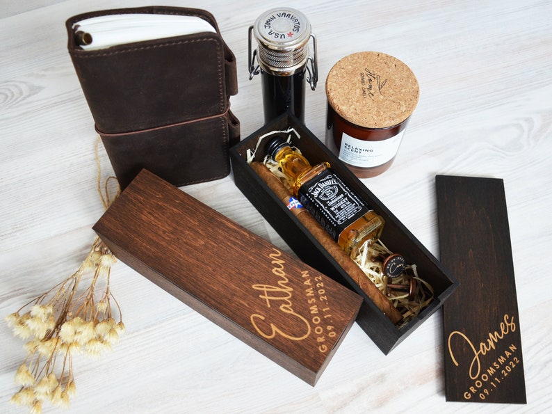 May include: A selection of gifts, including a brown leather journal, a small bottle with a metal clasp, a candle, and a wooden box containing a bottle of Jack Daniel's whiskey and a cigar. The box is engraved with the name "Ethan" and the date "09.11.2022".