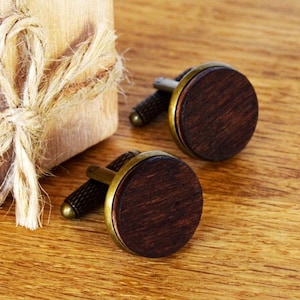 Puede incluir: Un par de gemelos redondos con incrustaciones de madera marrón oscuro en marcos de metal color latón antiguo. Los gemelos tienen un poste cilíndrico texturizado y un cierre redondeado, mostrando un acabado de veta de madera natural en la cara.