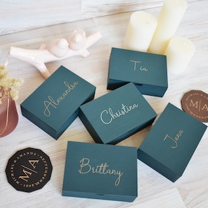 May include: Teal wooden boxes with gold lettering, each inscribed with a name: Alexandra, Christina, Brittany, Jena, and Tia. Two coasters with initials and a date are visible, alongside candles and decorative items.