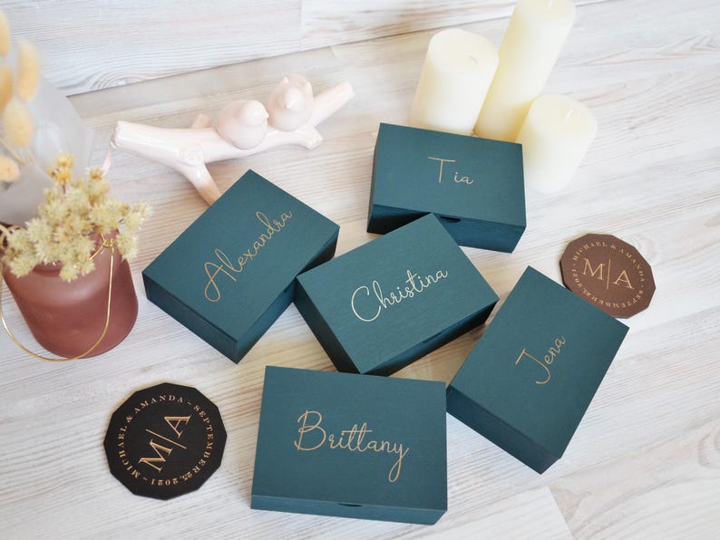 May include: Teal wooden boxes with gold lettering, each inscribed with a name: Alexandra, Christina, Brittany, Jena, and Tia. Two coasters with initials and a date are visible, alongside candles and decorative items.
