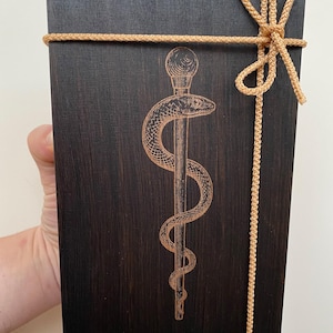 May include: A dark stained wooden box with a gold-coloured rope tied around it. The box features an engraved caduceus symbol, a staff with two snakes. The rope is tied in a decorative bow at the top.