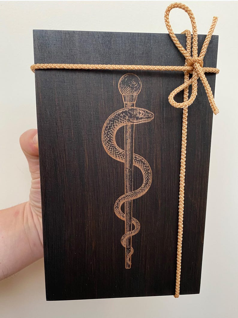 May include: A dark stained wooden box with a gold-coloured rope tied around it. The box features an engraved caduceus symbol, a staff with two snakes. The rope is tied in a decorative bow at the top.