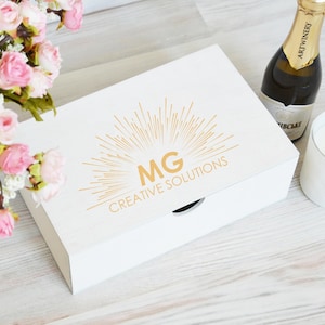 May include: A white box with gold text reading "MG CREATIVE SOLUTIONS", alongside a champagne bottle and a white candle. Pink and white roses are visible. The box has a circular opening.