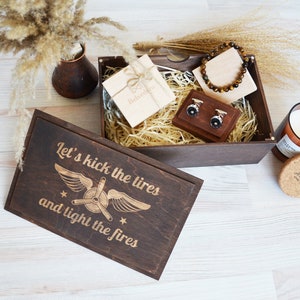 May include: A wooden gift box with a brown lid and a brown base. The lid has a design of a plane with the text "Let's kick the tires and light the fires". The box is filled with brown packing material and contains a small gift box with a brown ribbon, a pair of cufflinks, and a brown bracelet.