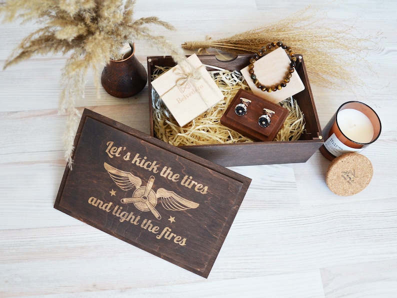 May include: A wooden gift box with a brown lid and a brown base. The lid has a design of a plane with the text "Let's kick the tires and light the fires". The box is filled with brown packing material and contains a small gift box with a brown ribbon, a pair of cufflinks, and a brown bracelet.
