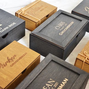 May include: Several wooden boxes in shades of brown and grey, some with engraved names and text. The rectangular boxes have hinged lids. Some are tied with twine. One box features the text "UP AND JOIN THE DARK SIDE".