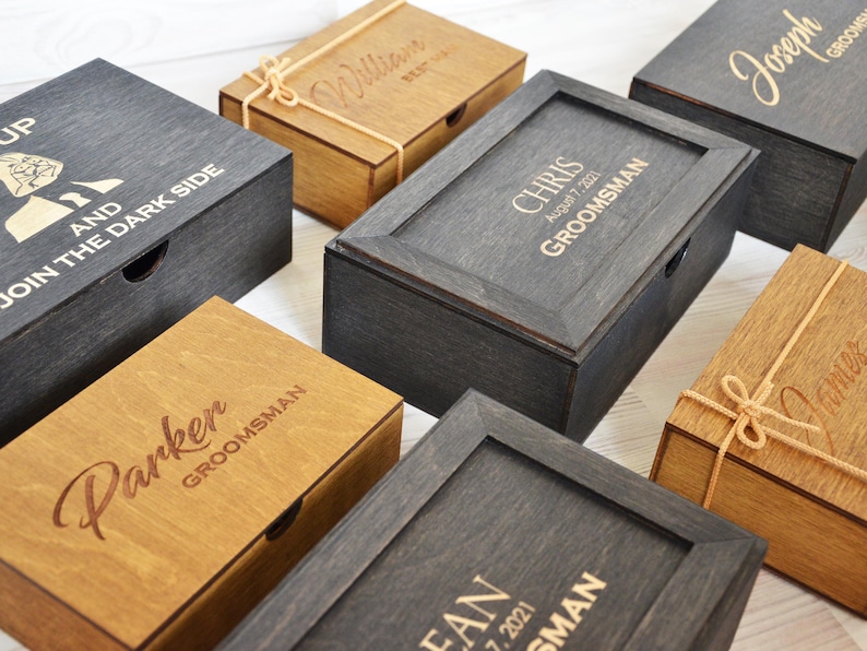 May include: Several wooden boxes in shades of brown and grey, some with engraved names and text. The rectangular boxes have hinged lids. Some are tied with twine. One box features the text "UP AND JOIN THE DARK SIDE".