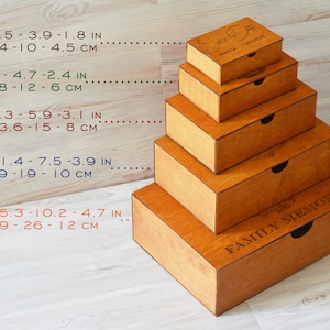 May include: Set of five wooden boxes with lids, ranging in size from extra small to extra large. The boxes are stacked on top of each other, with the largest box at the bottom. The boxes are all made of natural wood and have a light brown finish. The lids of the boxes are engraved with the words "Family Memories". The boxes are labeled with their size and dimensions in inches and centimeters.