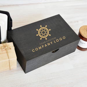 Pode incluir: Uma caixa de presente de madeira preta com um logotipo dourado de roda de navio e o texto "YOUR CUSTOM COMPANY LOGO". A caixa está aberta e contém uma pequena caixa de madeira com um laço de corda marrom e um frasco de vidro com uma tampa de cortiça.