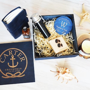 May include: A navy blue gift set with nautical elements. It features a personalised wooden box with an anchor, a leather notebook, a small bottle, a coaster, a candle, and a bottle with a swing top, all arranged in a wooden box.