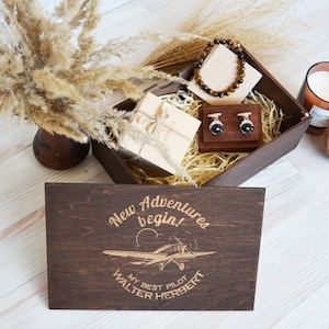 May include: A wooden box with a lid that has the text "New Adventures begin!" and "My Best Pilot Walter Herbert" engraved on it. The box contains a brown bracelet, cufflinks, a small wooden box, and a candle.