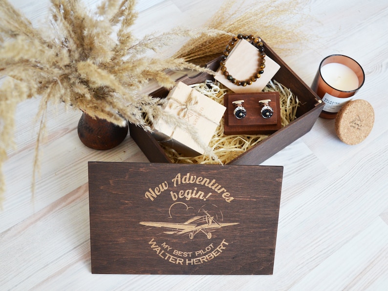 May include: A wooden box with a lid that has the text "New Adventures begin!" and "My Best Pilot Walter Herbert" engraved on it. The box contains a brown bracelet, cufflinks, a small wooden box, and a candle.