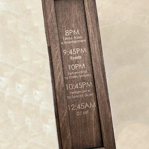 May include: A dark brown wooden sign with a rectangular frame. The sign displays event details, including times and activities such as "Drinks, Food & Entertainment," "Toasts," and "DJ set."