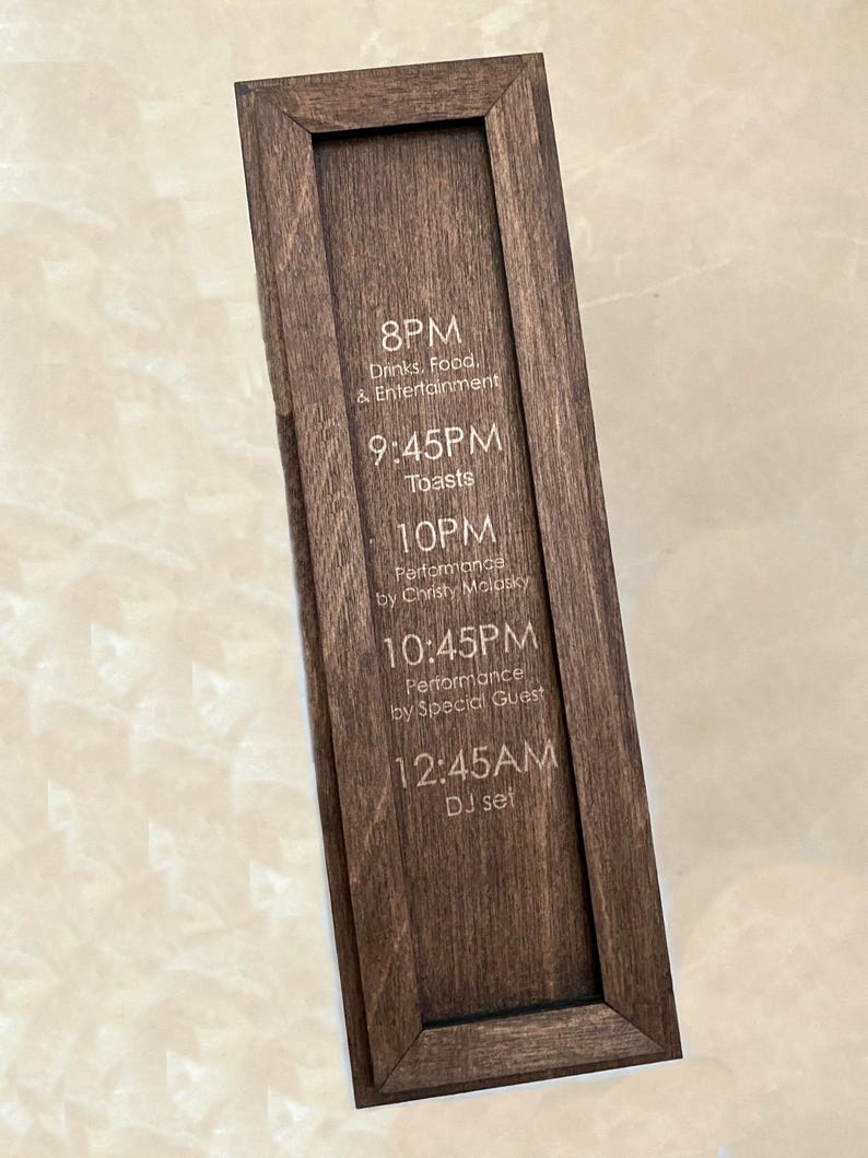 May include: A dark brown wooden sign with a rectangular frame. The sign displays event details, including times and activities such as "Drinks, Food & Entertainment," "Toasts," and "DJ set."