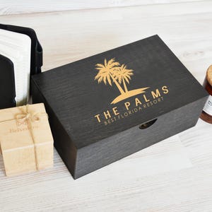 May include: A black wooden box with gold text and a palm tree logo that reads "THE PALMS BEST FLORIDA RESORT". A small wooden box tied with twine, a black leather notepad holder, and a brown candle with a cork lid are also present.