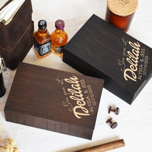 May include: Two wooden boxes with engraved text reading "See you at Delilah, Alyssa & Jess 07/15/2023". The boxes are dark brown and black. Also shown are miniature liquor bottles, a leather journal, a jar, and cufflinks.