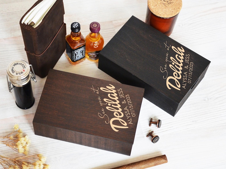 May include: Two wooden boxes with engraved text reading "See you at Delilah, Alyssa & Jess 07/15/2023". The boxes are dark brown and black. Also shown are miniature liquor bottles, a leather journal, a jar, and cufflinks.
