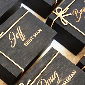 May include: A collection of black wooden gift boxes, each adorned with gold lettering and tied with gold twine. The boxes are inscribed with names and titles, including "Mike GROOMSMAN" and "Jeff BEST MAN", suggesting they are for a wedding.