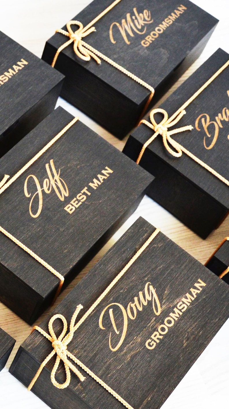 May include: A collection of black wooden gift boxes, each adorned with gold lettering and tied with gold twine. The boxes are inscribed with names and titles, including "Mike GROOMSMAN" and "Jeff BEST MAN", suggesting they are for a wedding.