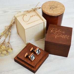May include: A selection of wooden gift boxes and accessories. Features a light-coloured box with "Conor GROOMSMAN" engraved, a dark wooden box with a name, a candle, and cufflinks with a black and white pattern.