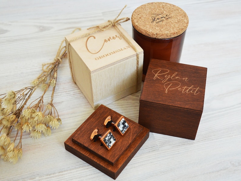 May include: A selection of wooden gift boxes and accessories. Features a light-coloured box with "Conor GROOMSMAN" engraved, a dark wooden box with a name, a candle, and cufflinks with a black and white pattern.