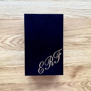 May include: A navy blue rectangular box with the engraved initials "E R F" in a cursive font on the lower right corner. The box is set against a light brown wooden surface.
