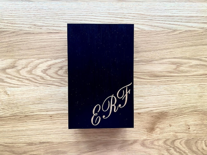 May include: A navy blue rectangular box with the engraved initials "E R F" in a cursive font on the lower right corner. The box is set against a light brown wooden surface.