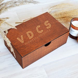 May include: A brown wooden box with the engraved text "VDCS MIDWEST LOGISTICS" on the lid. A candle in a glass jar and a cork coaster are next to the box. Dried plants are in the background. The box is a warm brown colour.