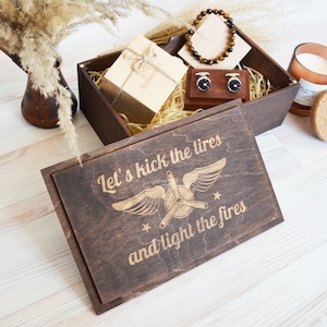May include: A wooden box with a lid that has an engraved design of an airplane with wings and the text "Let's kick the tires and light the fires". The box is filled with brown shredded paper and contains a small gift box, a pair of cufflinks, a bracelet, and a candle.