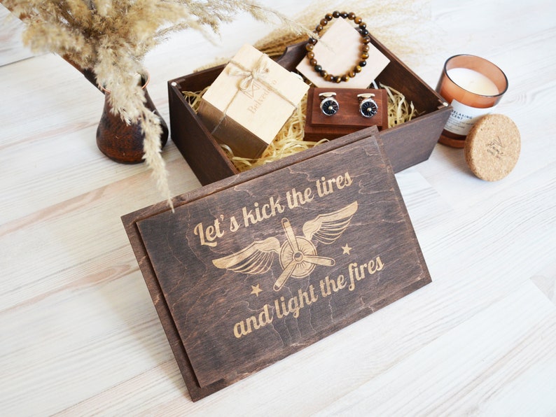May include: A wooden box with a lid that has an engraved design of an airplane with wings and the text "Let's kick the tires and light the fires". The box is filled with brown shredded paper and contains a small gift box, a pair of cufflinks, a bracelet, and a candle.