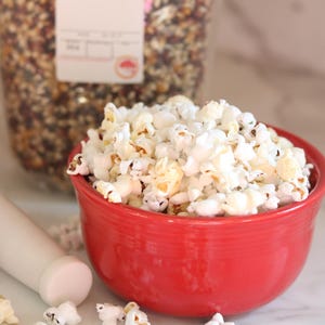May include: A red bowl filled with white popcorn. The popcorn is scattered on a white surface. A bag of multi-colored popcorn kernels is in the background.