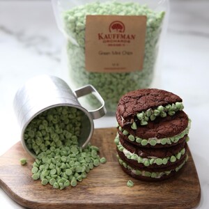 May include: A stack of four chocolate chip cookies with green mint chips and white frosting. A metal cup with green mint chips is spilling onto a wooden cutting board. A bag of green mint chips with the label "Kauffman Orchards Green Mint Chips" is in the background.