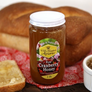 May include: A glass jar of raw cranberry honey with a white lid. The jar has a label that says "Pure & Natural Welsh Mountain Apiaries Raw Cranberry Honey 1 lb."