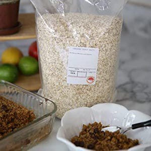 May include: A clear plastic bag filled with rolled oats. The bag has a white label with the text "Organic Quick Oats" printed on it. A glass baking dish filled with a brown mixture of oats and chocolate chips is in the foreground.