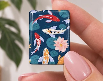 May include: A rectangular tile featuring a dark blue background with a pattern of swimming koi fish in red, white, and orange, interspersed with green lily pads and a single light pink lotus flower. The tile has smooth, rounded edges.