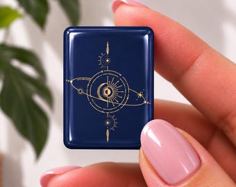 May include: A small, rectangular, dark navy blue tile featuring a gold-coloured celestial graphic. The design includes a central sun and moon motif surrounded by orbital rings, stars, and radiating lines, creating a detailed, symmetrical cosmic pattern.