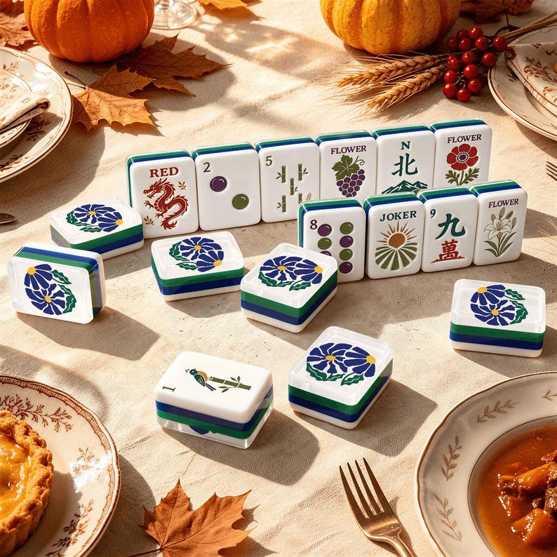 May include: A set of white mahjong tiles with green and blue edges, featuring various colorful illustrations including a red dragon, flowers, bamboo, and text such as RED, FLOWER, N, and JOKER, arranged on a light-colored surface with autumn decor.