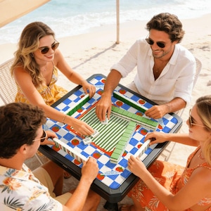 May include: A group of four people playing Mahjong at a table on a beach. The table has a blue and white checkered pattern with red tomato illustrations. The Mahjong tiles are white, green, and red.