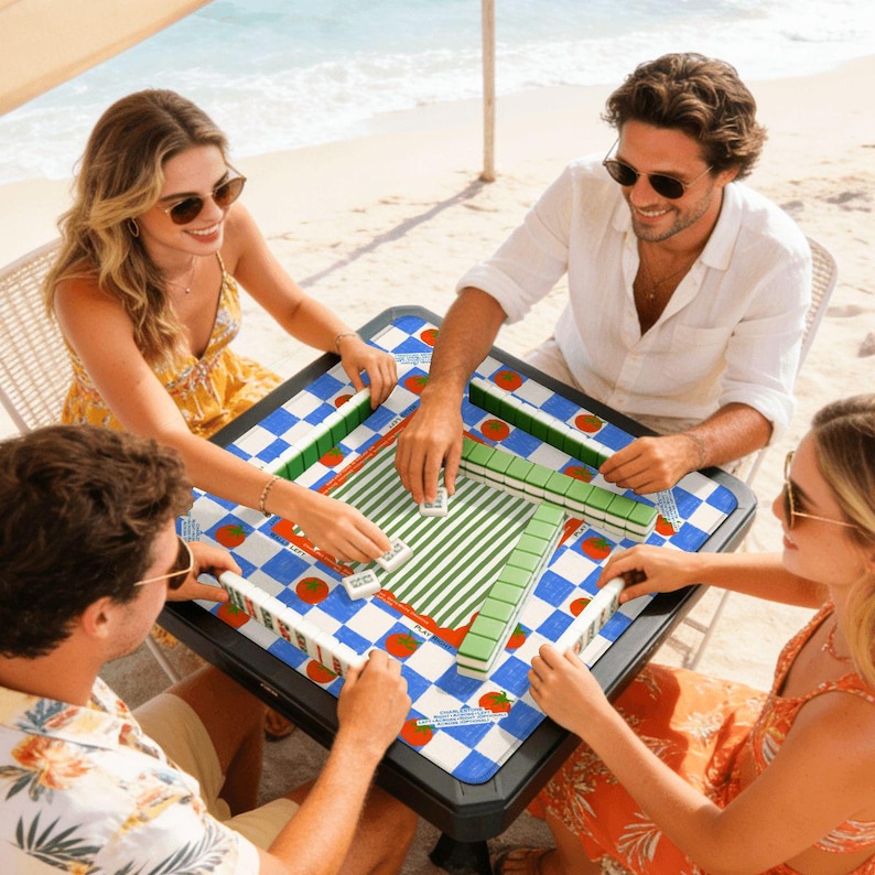 May include: A group of four people playing Mahjong at a table on a beach. The table has a blue and white checkered pattern with red tomato illustrations. The Mahjong tiles are white, green, and red.