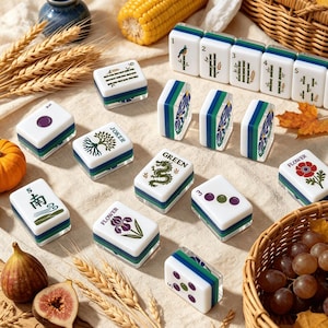 May include: A collection of white Mahjong tiles featuring various colorful illustrations including bamboo stalks, flowers, a green dragon, and the word JOKER. The tiles have blue and green layered bases and are arranged on a textured fabric surface.