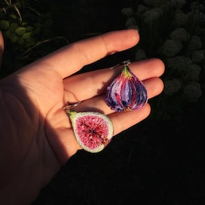 Könnte beinhalten: Zwei feigenförmige bestickte Anhänger mit komplexer Fadenarbeit. Ein Anhänger zeigt einen Feigenquerschnitt mit leuchtend rosa-rotem Inneren, während der andere die dunkelviolette und blaue Außenhaut einer ganzen Feige darstellt.