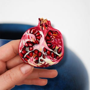 May include: A detailed, embroidered pomegranate half, showcasing vibrant red seeds and a textured rind. The embroidery captures the fruit's natural colors and intricate details, with a focus on the seeds and the inner white membrane.