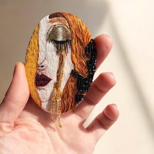 Könnte beinhalten: Oval geformte, gestickte Brosche mit einem stilisierten Gesicht mit geschlossenen Augen und fließendem Haar. Das Design verwendet Weiß-, Gold-, Rot- und schwarze Perlen. Das Gesicht hat eine tränenartige Verzierung mit hängenden Goldperlen.