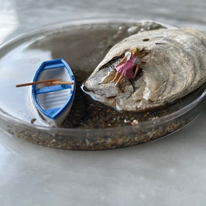 Może przedstawiać: Miniaturowa scena z małą niebiesko-białą łódką i miniaturową postacią siedzącą na skale w szklanej misce wypełnionej piaskiem i wodą.