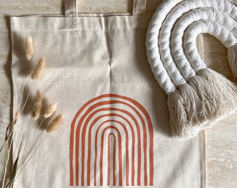 May include: A beige canvas tote bag with a red-orange rainbow design. The bag has long straps and is sitting on a light brown surface.