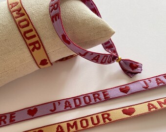 Op de afbeelding: Twee geweven armbanden met rode letters en kleine rode hartjes. Een armband is lavendelkleurig met de tekst "J'ADORE" en de andere is geel met de tekst "AMOUR".
