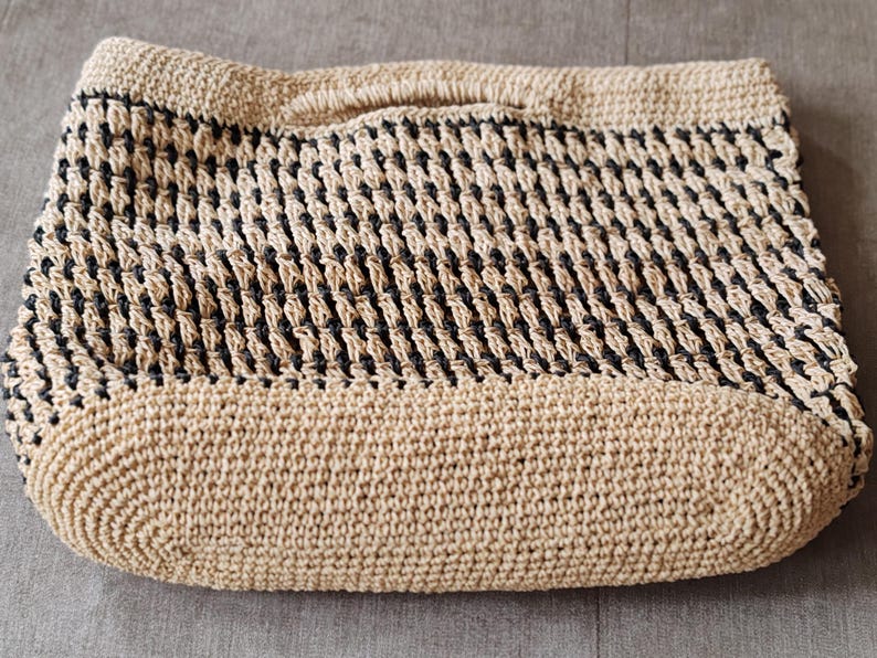 May include: A beige and black woven tote bag with a rectangular shape and a handle. The bag features a woven pattern with alternating beige and black sections. The bottom and top edges are solid beige.