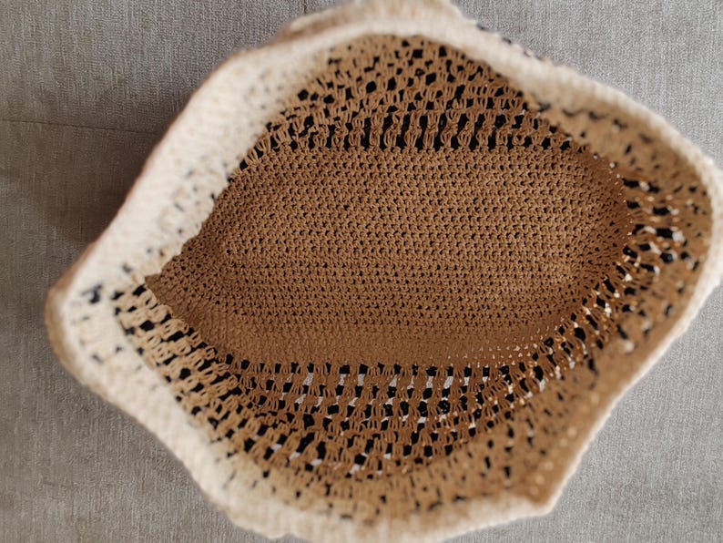 May include: A handmade crochet hat in a neutral color palette. The hat features a beige interior and a cream-colored brim. The open weave design allows for breathability, making it suitable for warm weather. The hat's shape is slightly rounded.