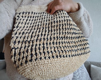 May include: A beige and black woven tote bag. The bag features a textured pattern with alternating rows of beige and black. The bag is held by a person wearing a beige sweater. The bag is a stylish accessory.